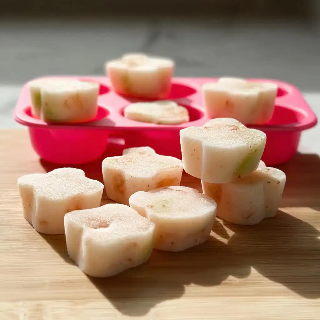 Several flower-shaped Frozen Apple Dog Treats for Summer resting on a wooden board next to a pink silicone mold.