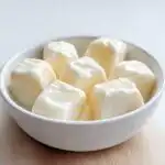 A white bowl filled with several creamy, pale yellow Frozen Yogurt Dog Cubes against a bright background.